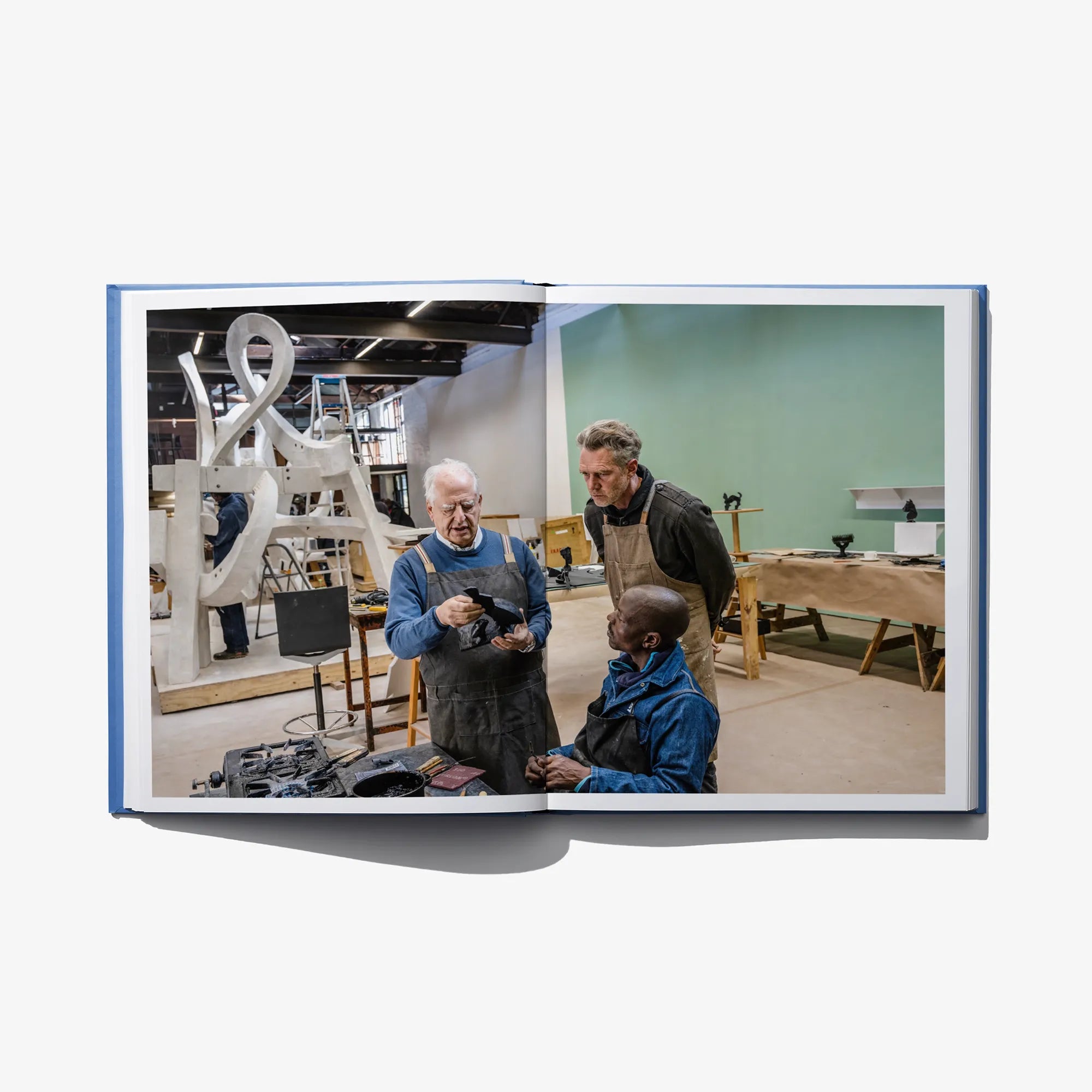 Open art book spread showing a workshop scene with people at work